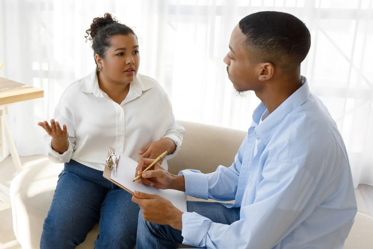 Young couple is sitting on a couch, discussing issues. The man is holding a clipboard listening to the woman speak.