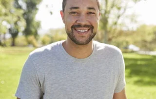 man looking at camera and smiling. he is wearing a grey tee shirt and is standing in front of trees