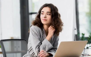 woman with dark hair sitting at an office desk in front of her laptop looking in the distance