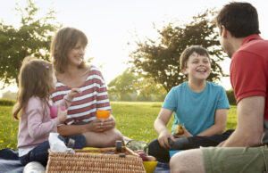a woman, man and two children sitting on the grass in a park taking part in a picnic.