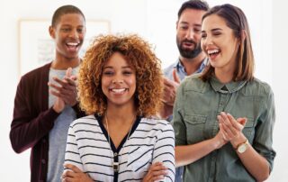 A woman smiling with arms crossed and three colleagues clapping for her showing appreciation for her.