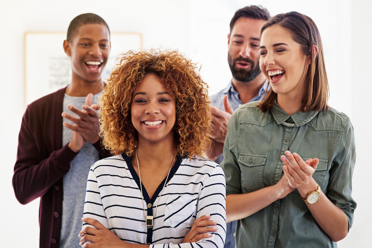 A woman smiling with arms crossed and three colleagues clapping for her showing appreciation for her.